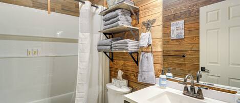 A bathroom with a white bathtub, a white toilet, a wooden wall, a shelf with towels, a sink, and a mirror.
