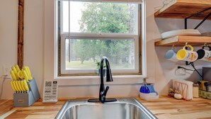 Una cocina con un lavamanos de acero inoxidable, un grifo negro, una ventana con vistas a árboles y un bloque de cuchillos en la encimera.