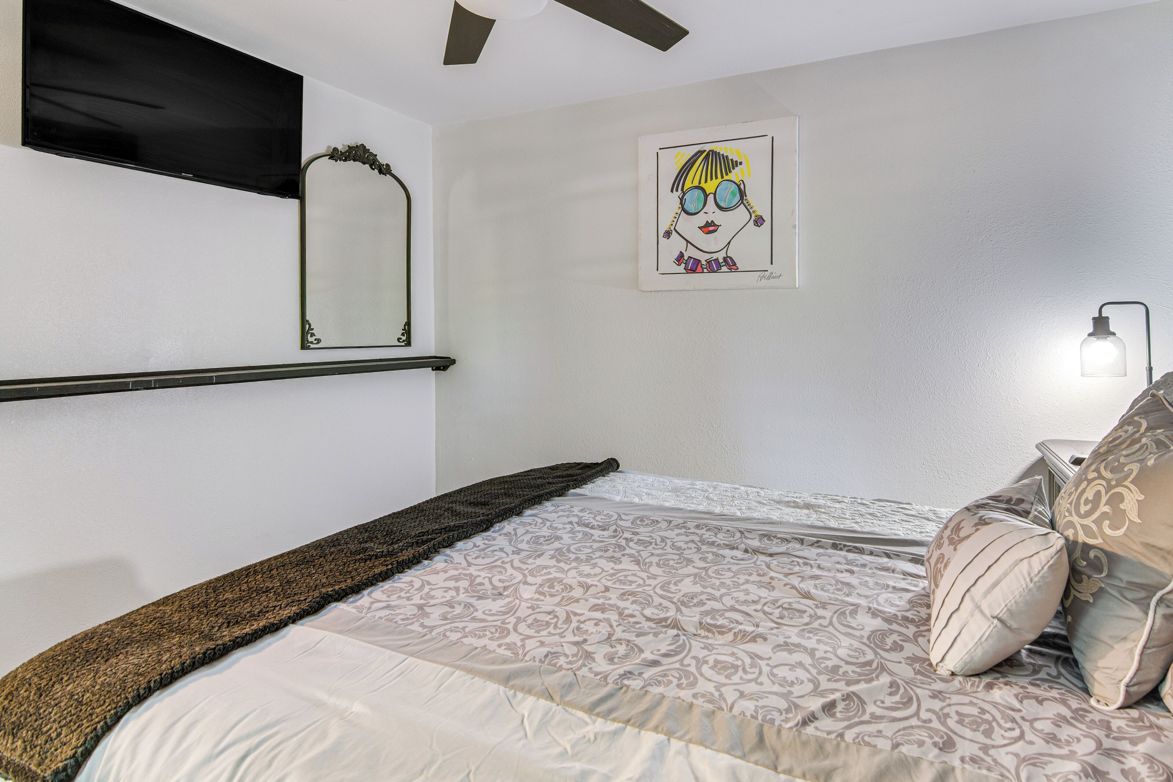 A bedroom with a bed, a TV mounted on the wall, a framed artwork, and a bedside lamp.
