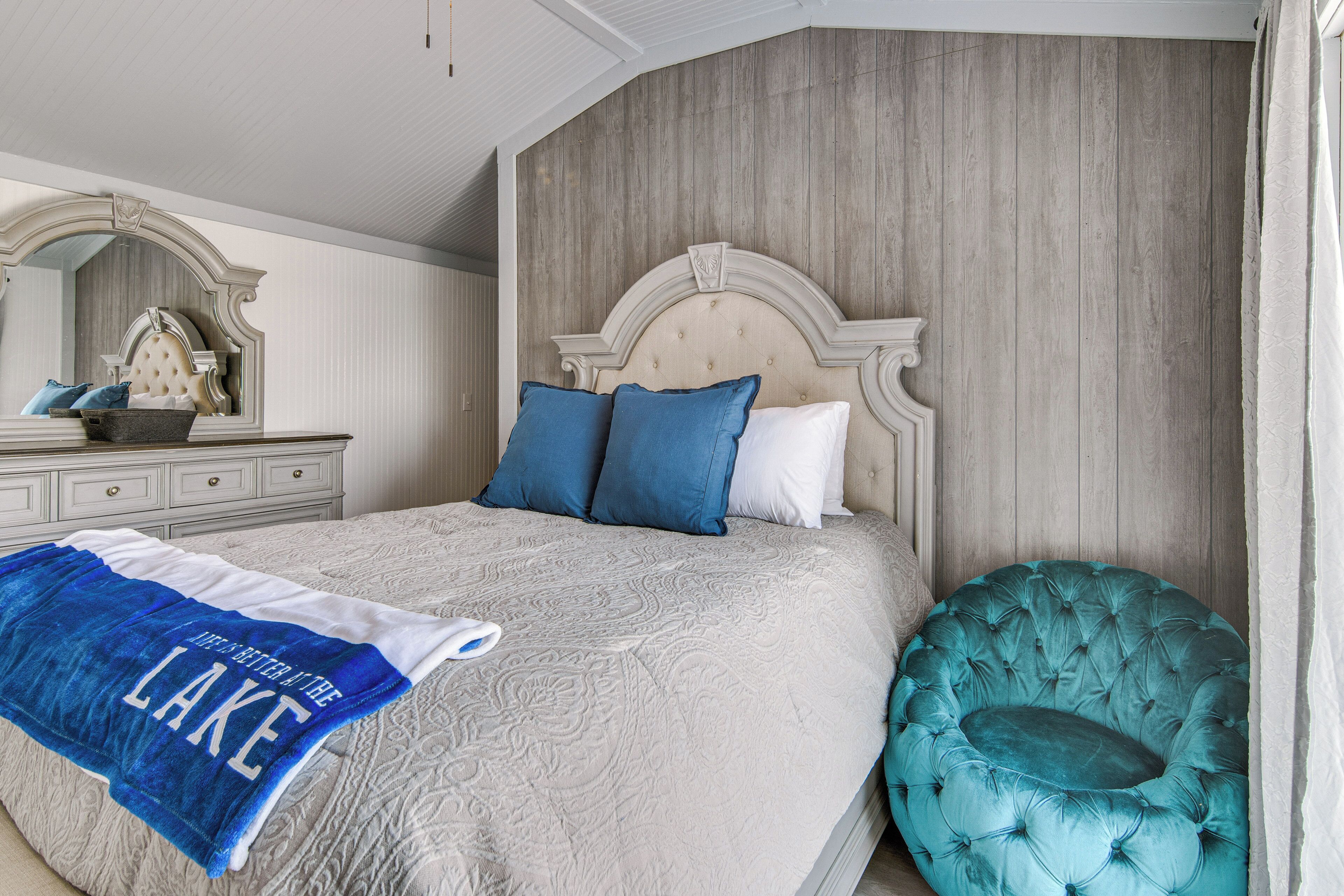 A bedroom with a large bed, a tufted headboard, a blue and white throw blanket, a round teal chair, and a wooden wall panel.