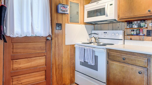 A compact kitchen with wooden cabinets, a white stove, a microwave, and a small shelf with kitchen essentials. - Dock & Rowboat: 'beechnut Bungalow' on Keuka Lake (Branchport)