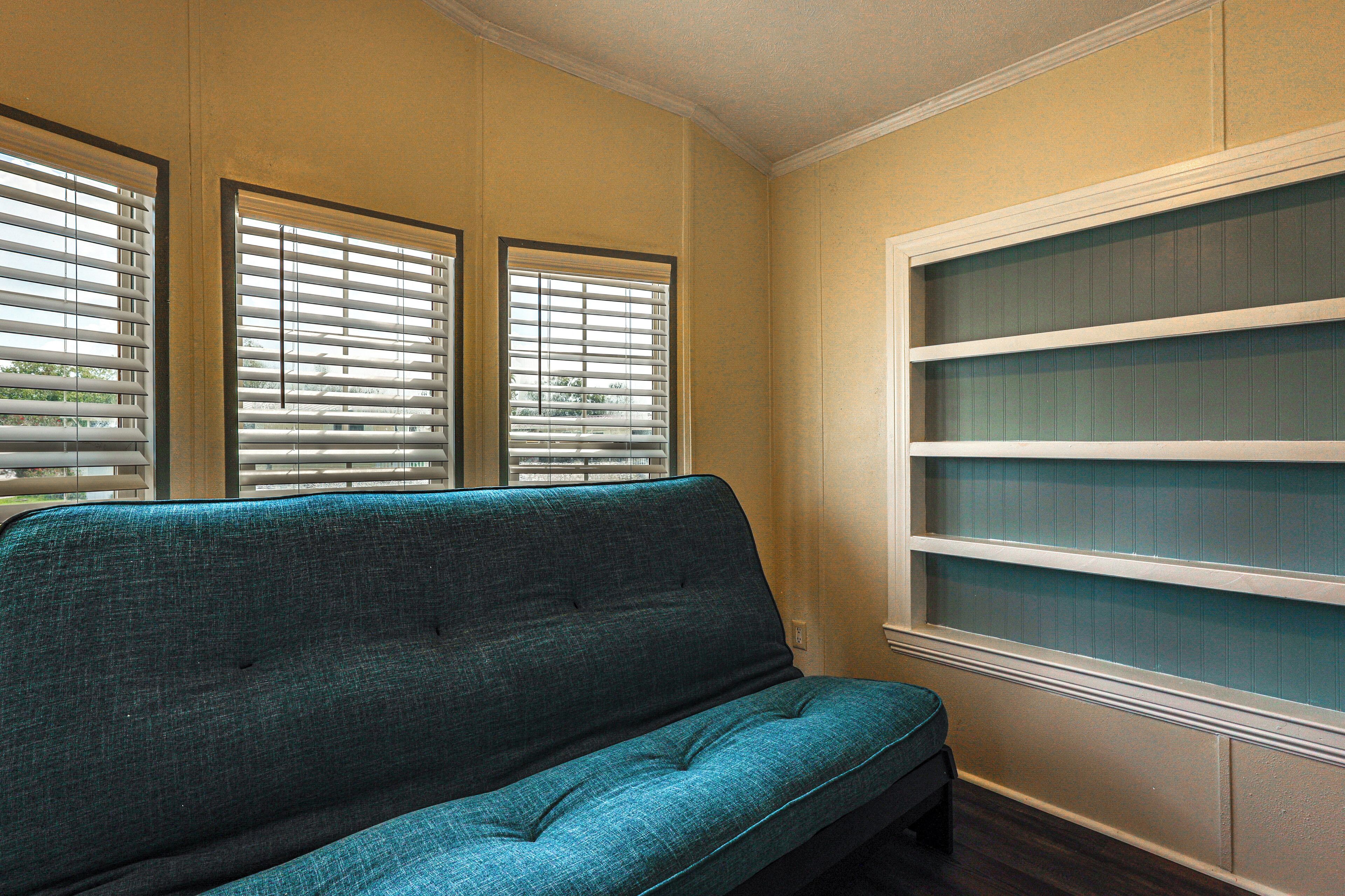 A room with a teal sofa, a window with blinds, and a built-in shelving unit.