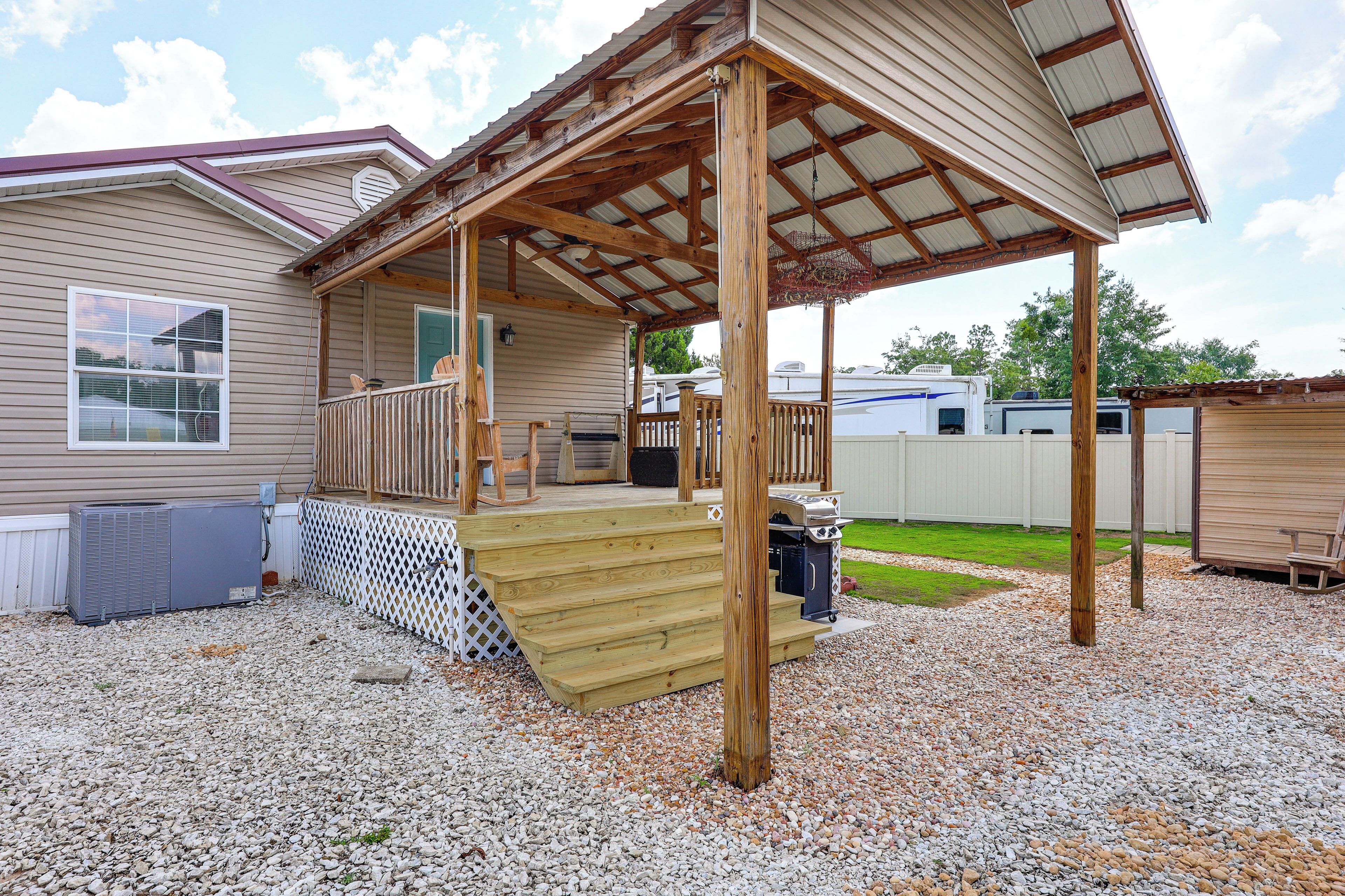 A covered porch with wooden steps leading to a deck, a grill, and a white fence in the background.