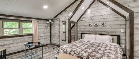 Un dormitorio con techo de madera, una cama con colcha estampada, un banco, un escritorio con computadora y un ventilador de techo.