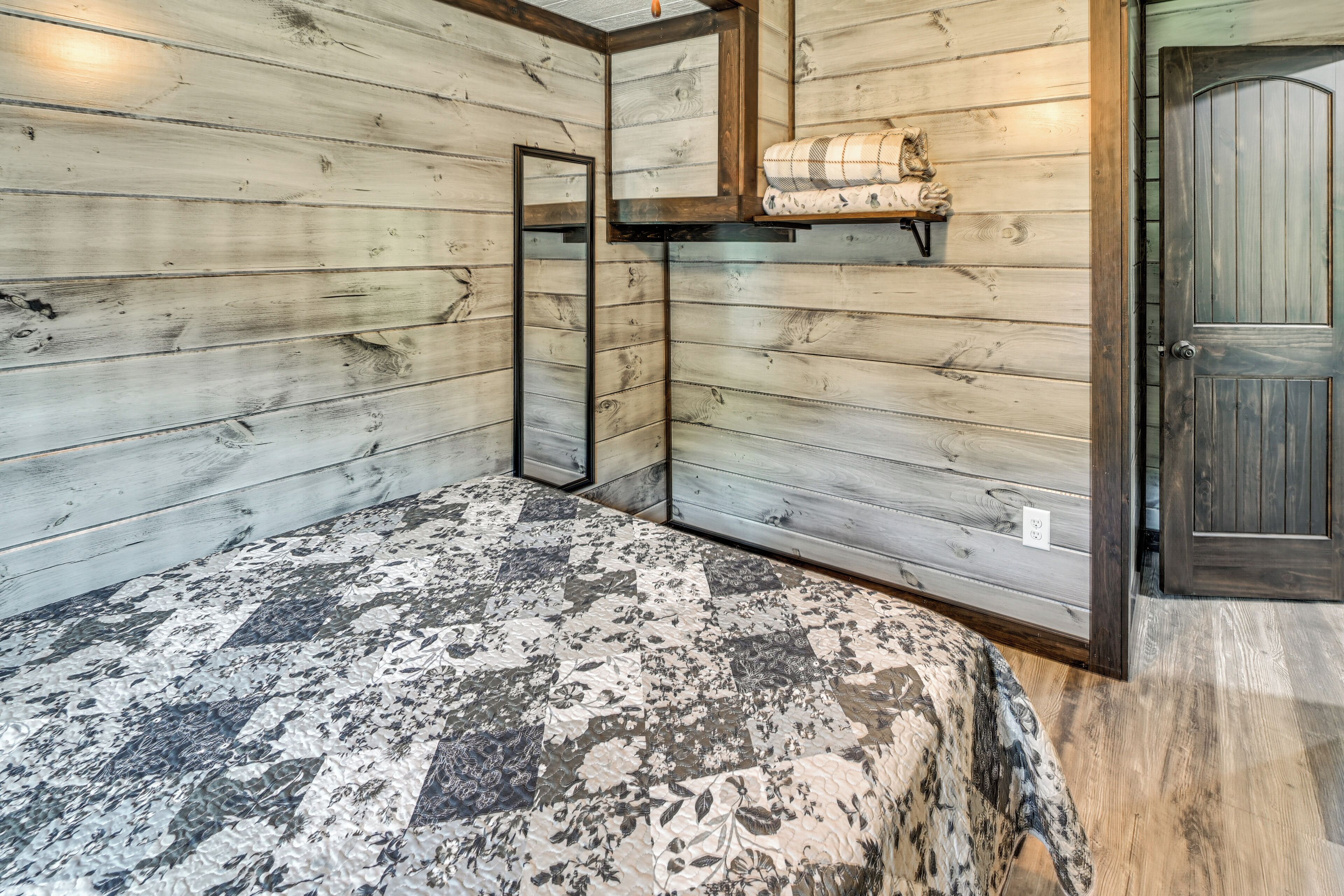 A room with wooden walls, a bed with a patterned bedspread, a mirror, and a shelf with a blanket.