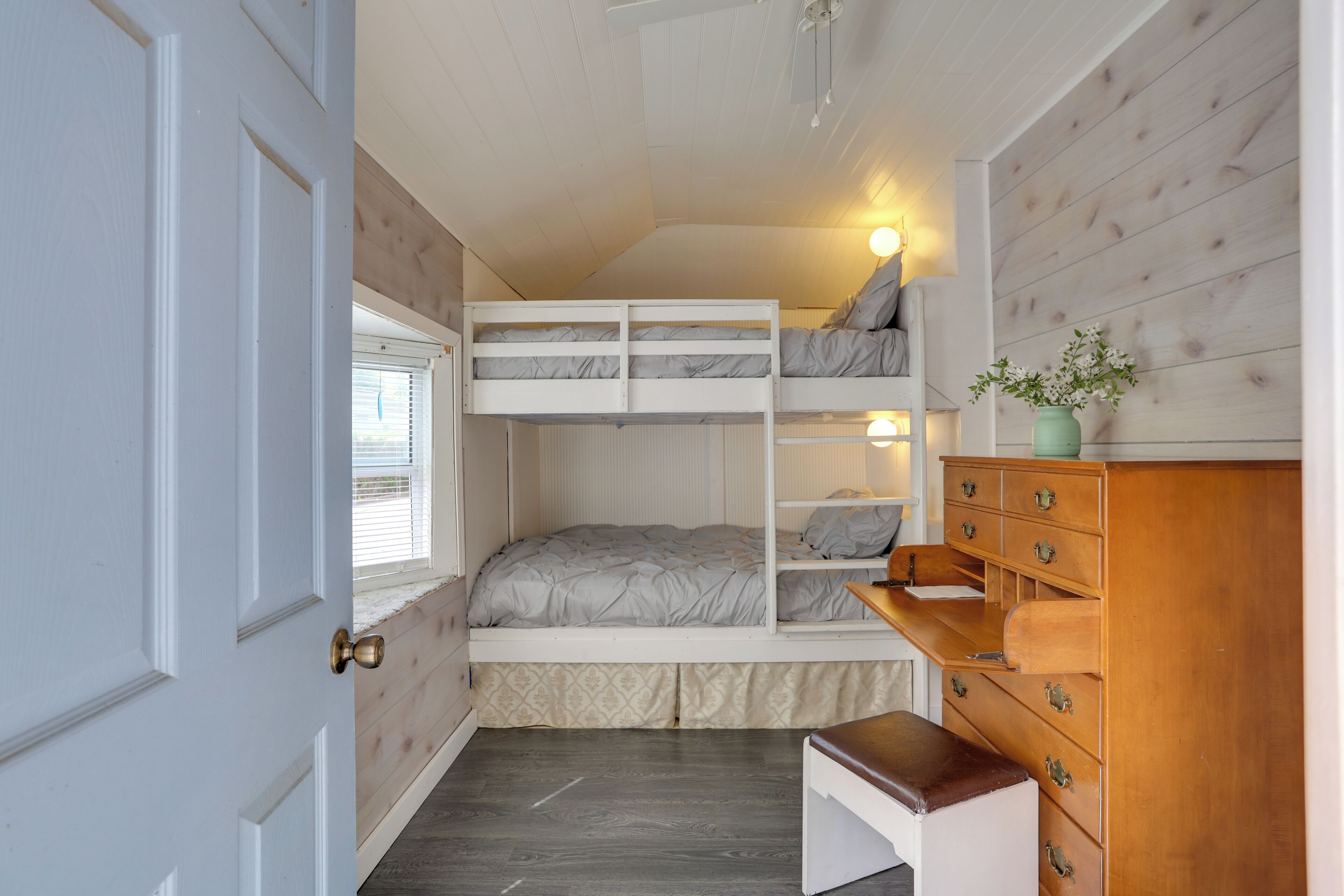 A bedroom with a bunk bed, a dresser, a bench, and a window with blinds.