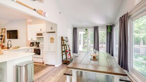 A modern kitchen with white cabinets, a wooden dining table, and a view of the outdoors through large windows.