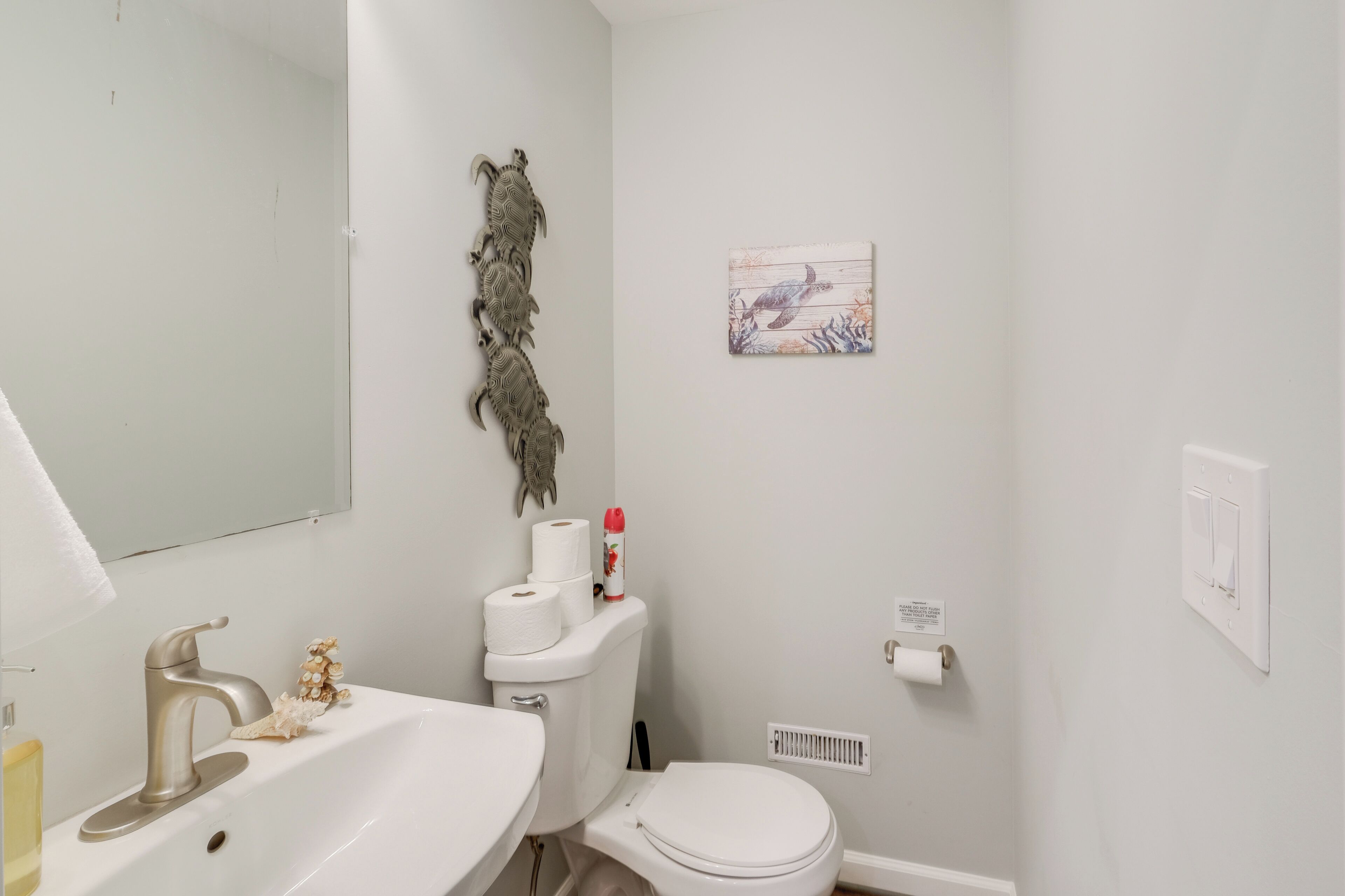 A bathroom with a white sink, toilet, and mirror. There is a decorative wall piece above the toilet and a framed picture on the wall.