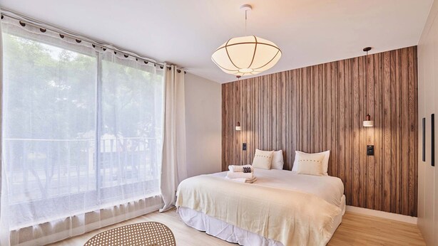 Un dormitorio moderno con una cama grande, un revestimiento de pared de madera, una zona de comedor con una mesa redonda y un ventanal con cortinas ligeras.