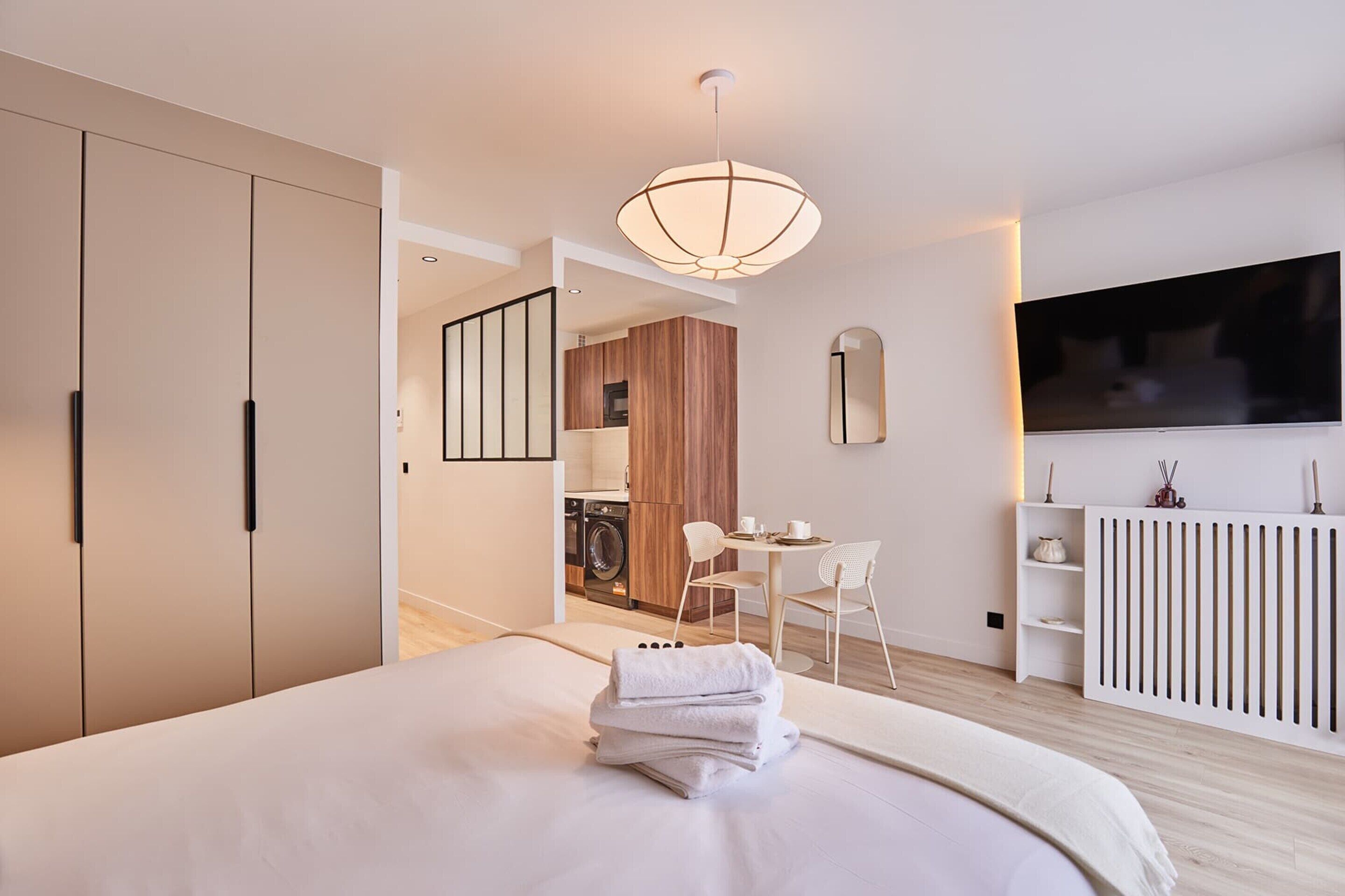 A modern hotel room with a large bed, a TV, a washing machine, and a dining area.