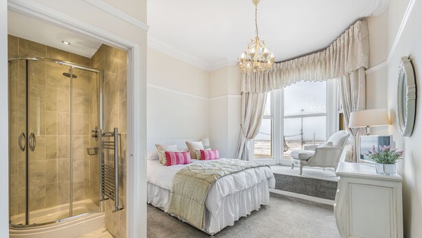 A spacious bedroom with a large bed, a walk-in shower, a sitting area with a chair and table, and a view of the outdoors. - Pier View House - Blackpool (Blackpool)
