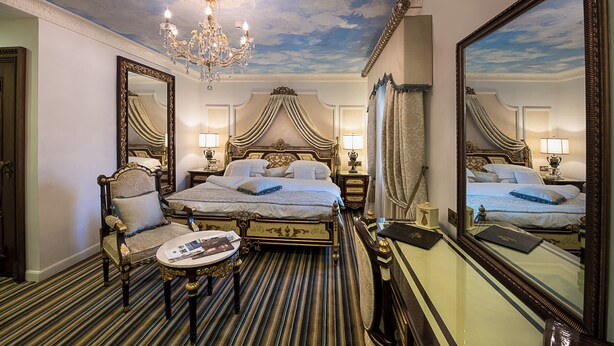 A luxurious hotel room with a large bed, a sitting area with a chair and a small table, a chandelier, and a large mirror reflecting the room. - House of splendor Boutique Hotel (Lagos)