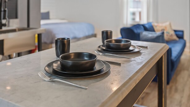 Een moderne eethoek met een marmeren tafel gedekt voor twee personen, een bed op de achtergrond en een blauwe bank met uitzicht op de stad.