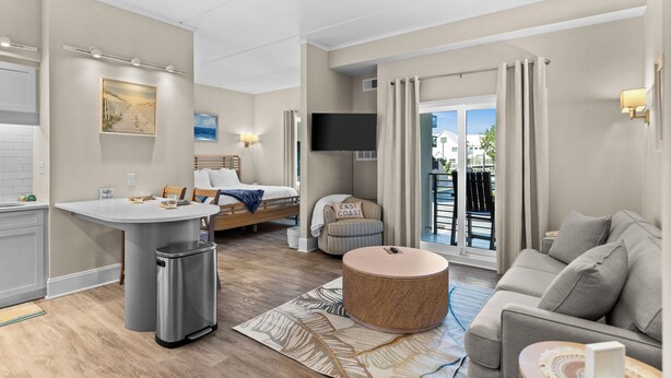 Una habitación de hotel moderna con kitchenette, una cama, zona de estar con sofá y reposapiés, televisión y balcón con cortinas.