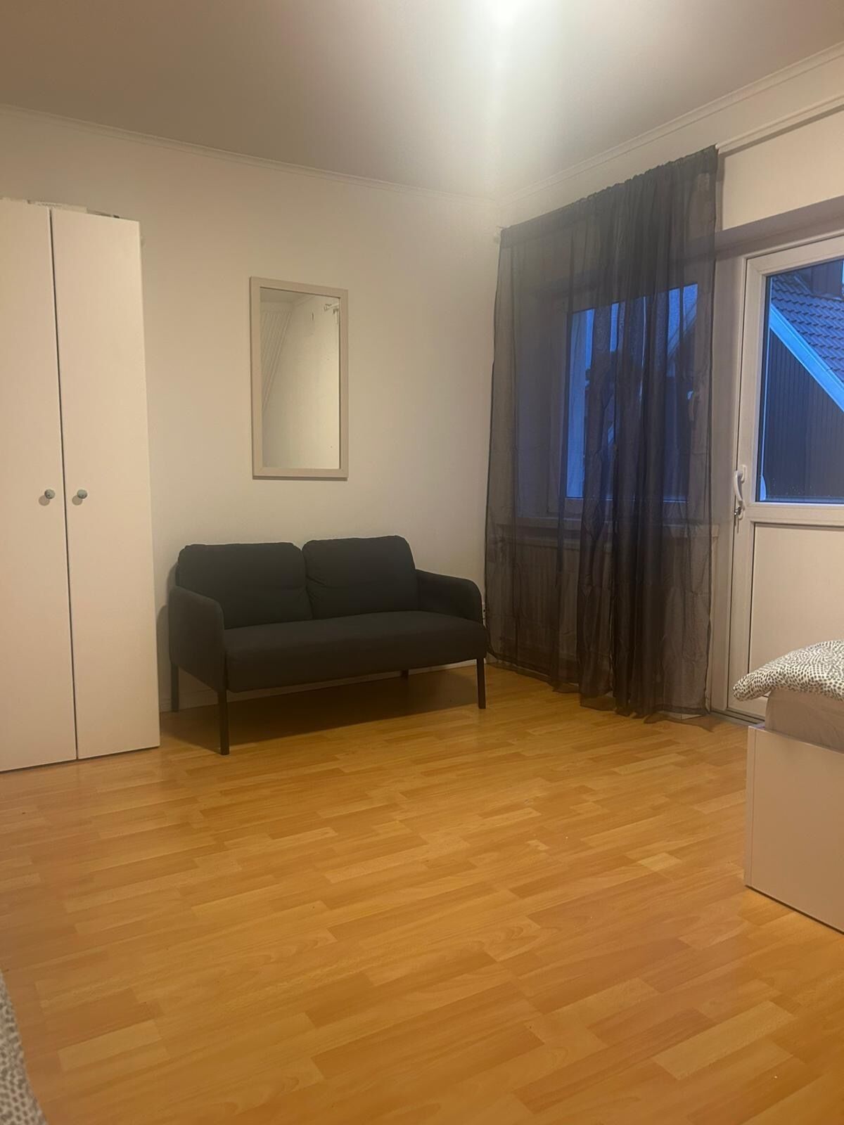 Una habitación con un sofá de color oscuro, un armario blanco, un cuadro enmarcado en la pared y una ventana con cortinas oscuras.