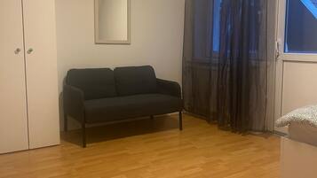 Una habitación con un sofá de color oscuro, un armario blanco, un cuadro enmarcado en la pared y una ventana con cortinas oscuras.