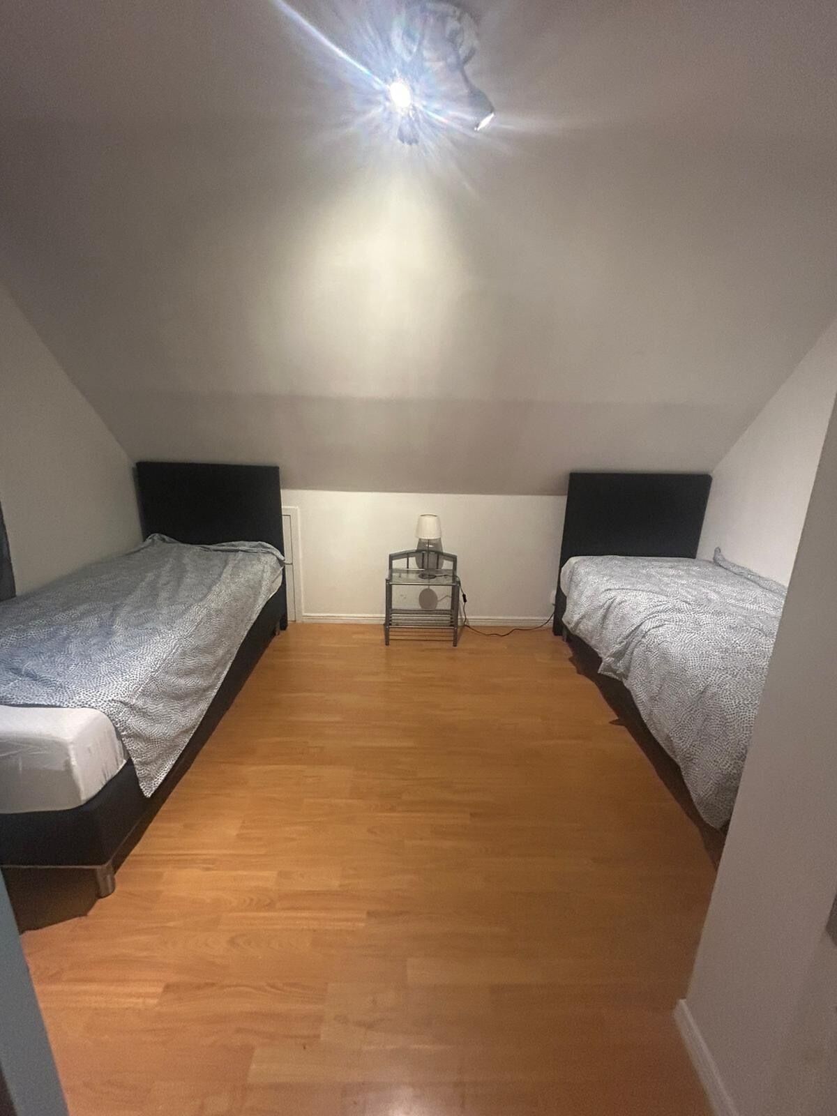 Une petite pièce faiblement éclairée, avec deux lits, une petite table et un luminaire au plafond.