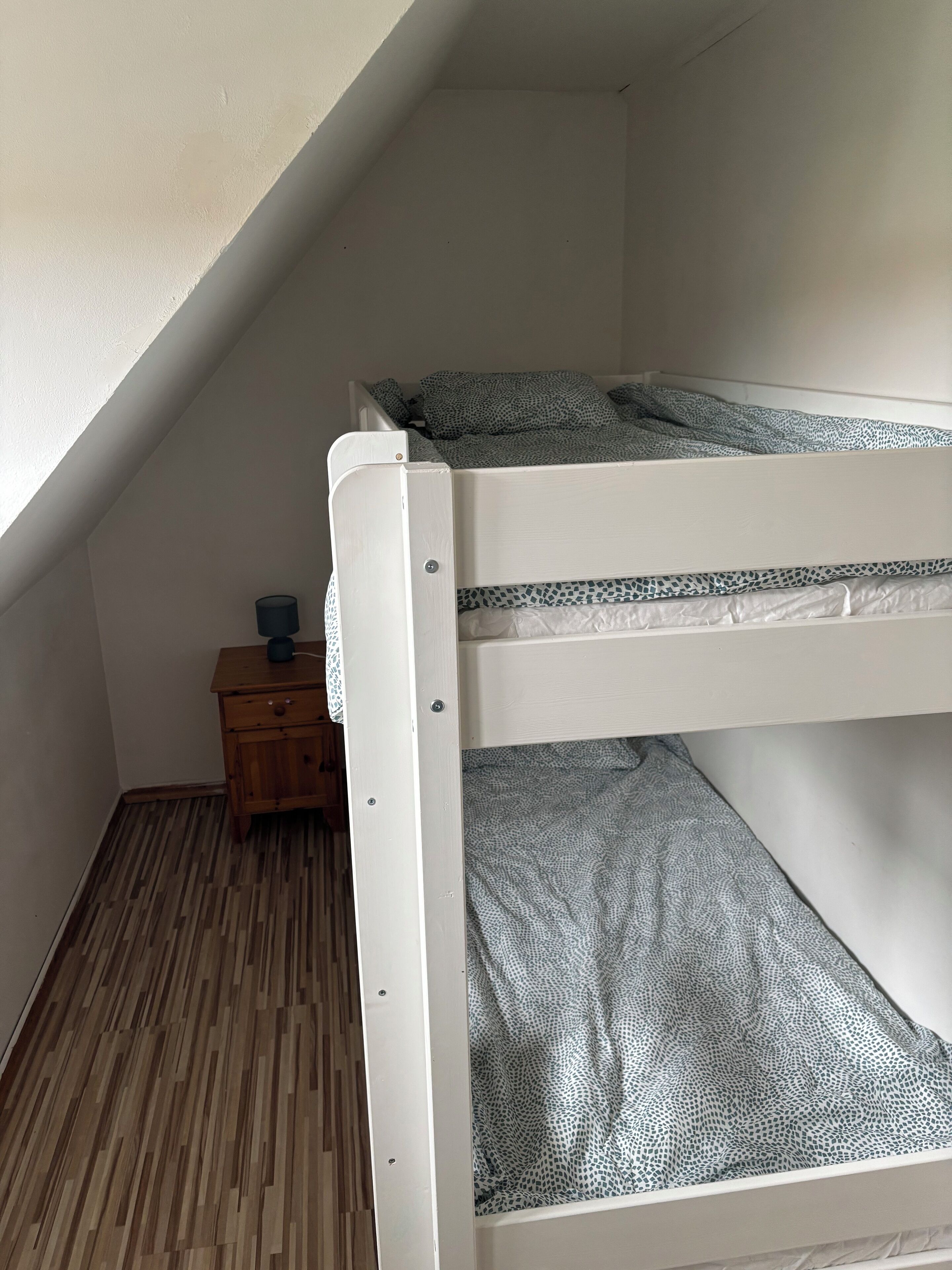 Una cama litera con ropa de cama estampada, una mesita de noche de madera con una lámpara y un techo inclinado.