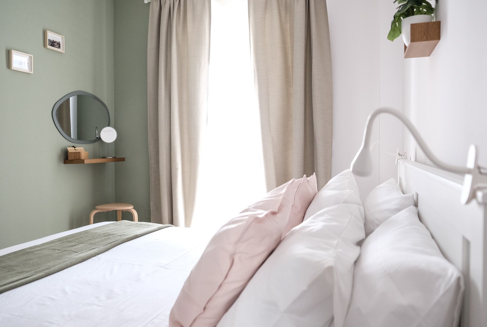 Un letto rifatto con lenzuola bianche e rosa, uno specchio rotondo su una mensola di legno e un piccolo sgabello.