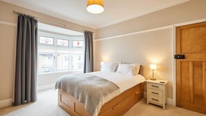 Ein Schlafzimmer mit einem hölzernen Bett, einem Nachttisch mit Lampe und Blick ins Freie durch ein Fenster mit Vorhängen.