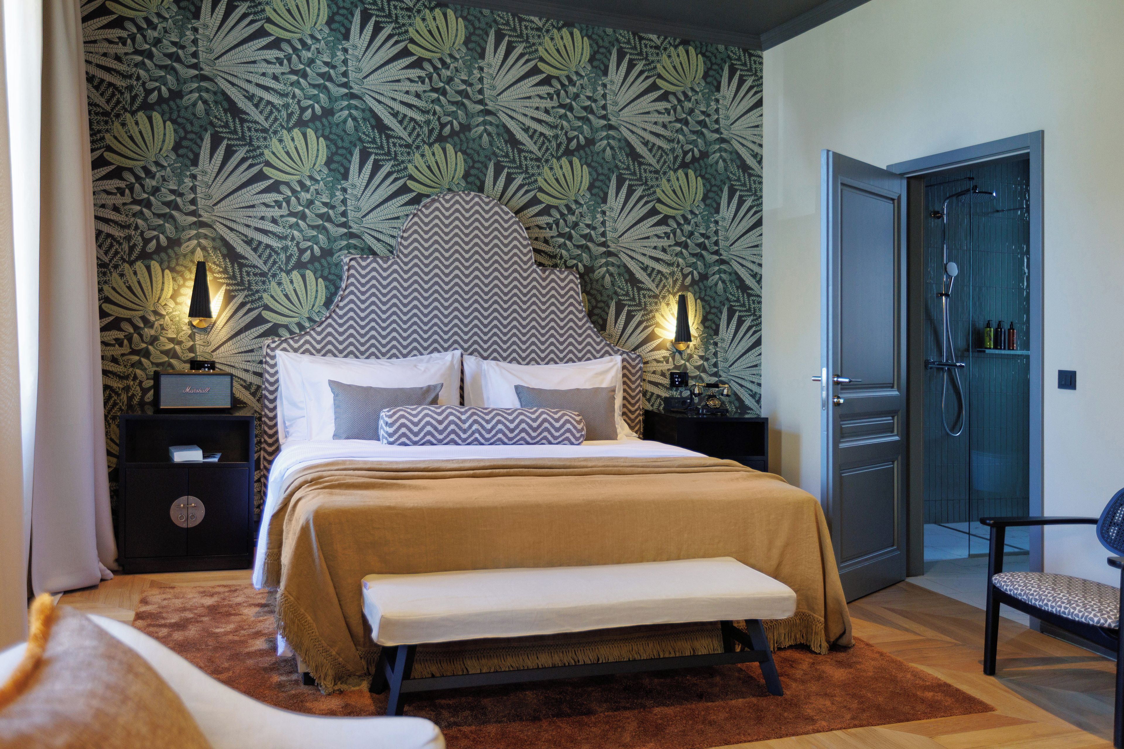 A bedroom with a large bed, a bench, a nightstand, and a wall with a leafy pattern.