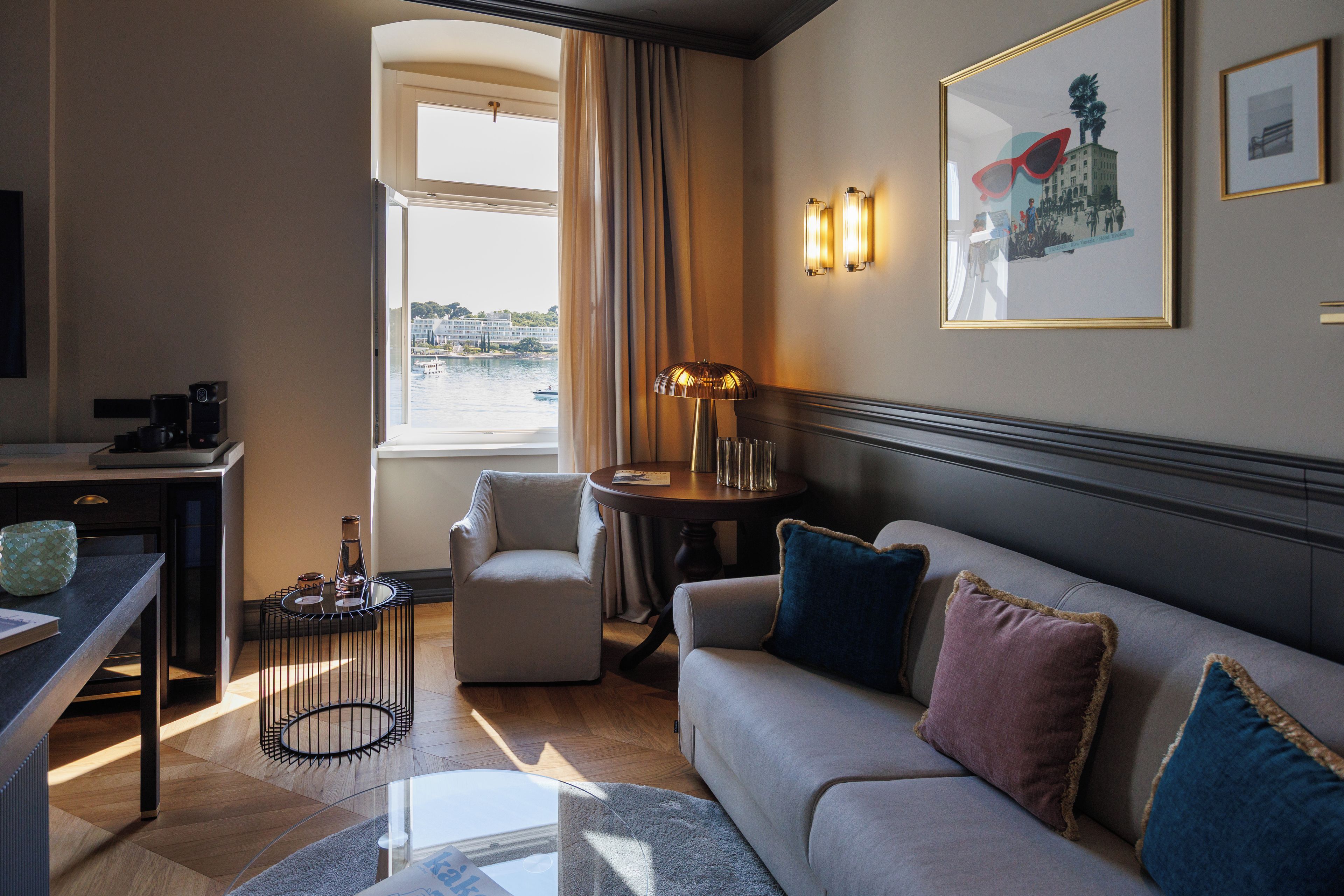 Habitación de hotel con sofá, sillón, mesa y vista a un cuerpo de agua a través de una ventana.