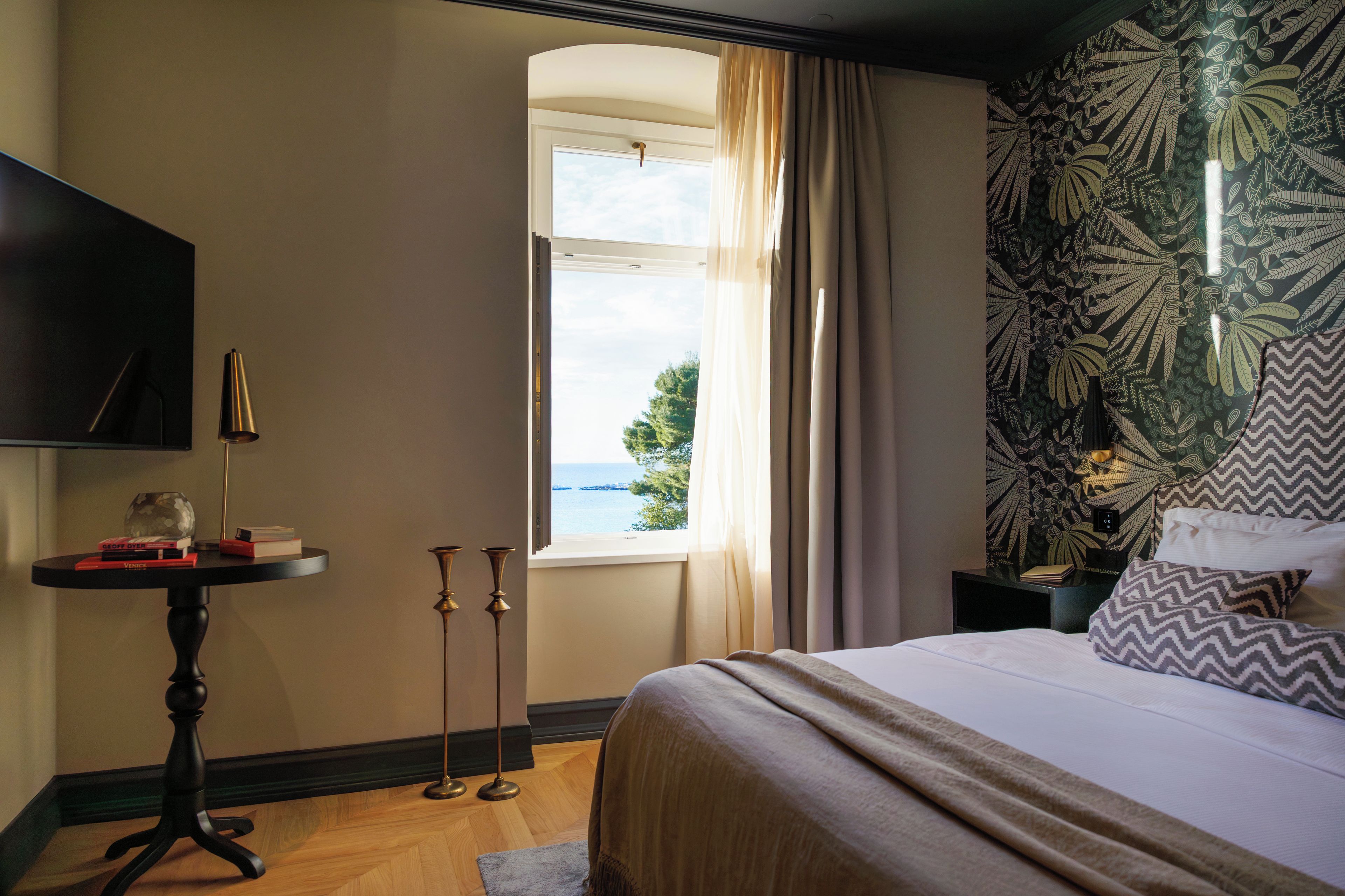 A bedroom with a bed, a table with books, a lamp, a window with curtains, and a patterned wall.
