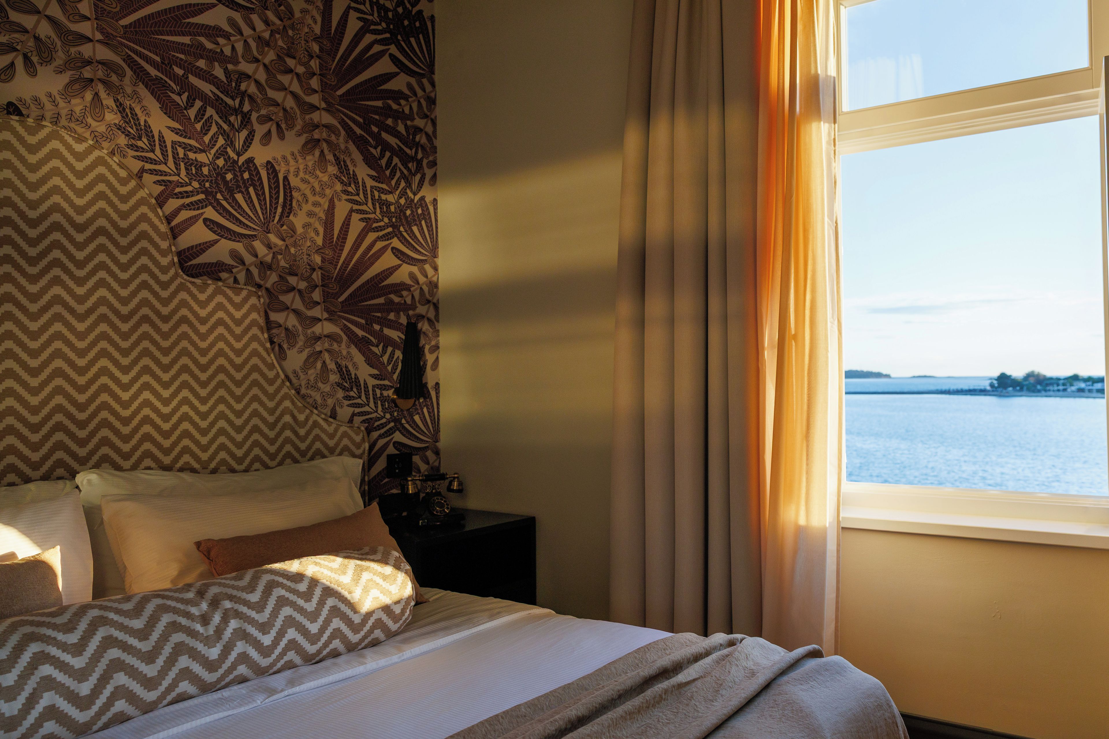 A hotel room with a bed, a nightstand, a lamp, and a window with a view of the water.