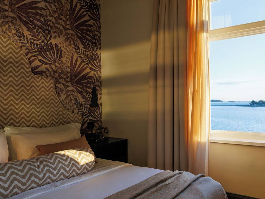 A hotel room with a bed, a nightstand, a lamp, and a window with a view of the water.