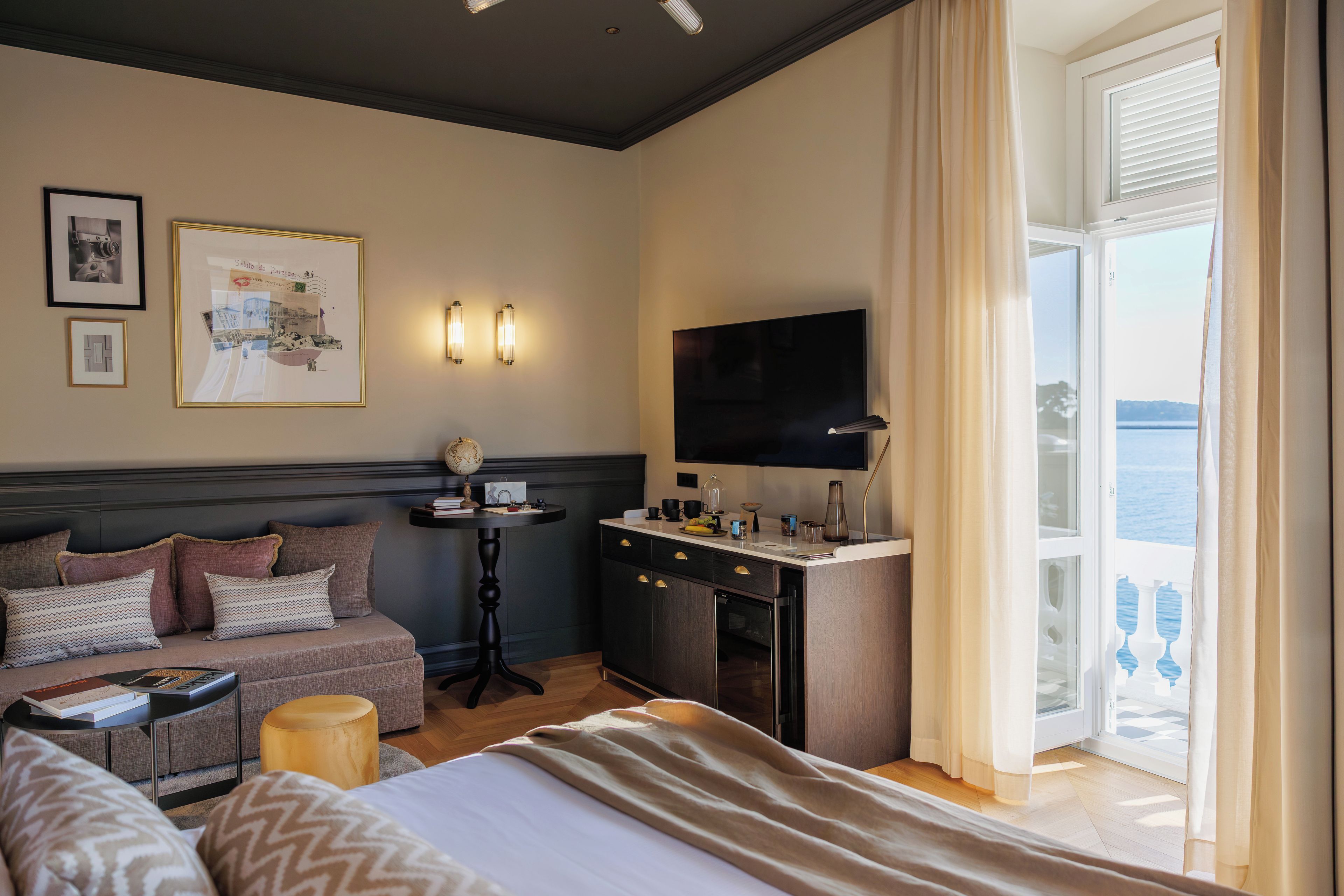 Ein Hotelzimmer mit Bett, einer Couch, einem Schreibtisch mit Flachbildfernseher, einem Globus und Meerblick durch ein großes Fenster.