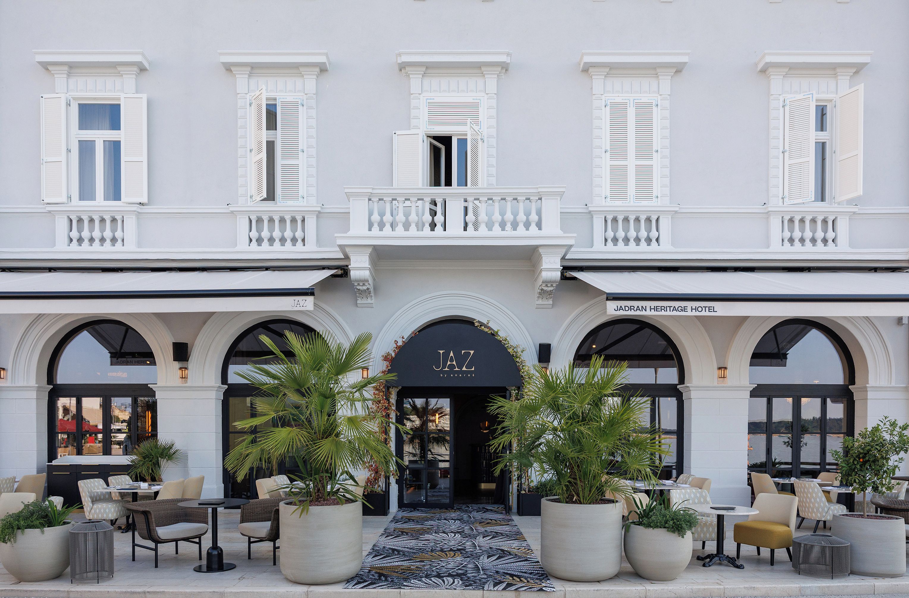 A hotel facade with a central entrance, arched windows, and outdoor seating area with potted plants.