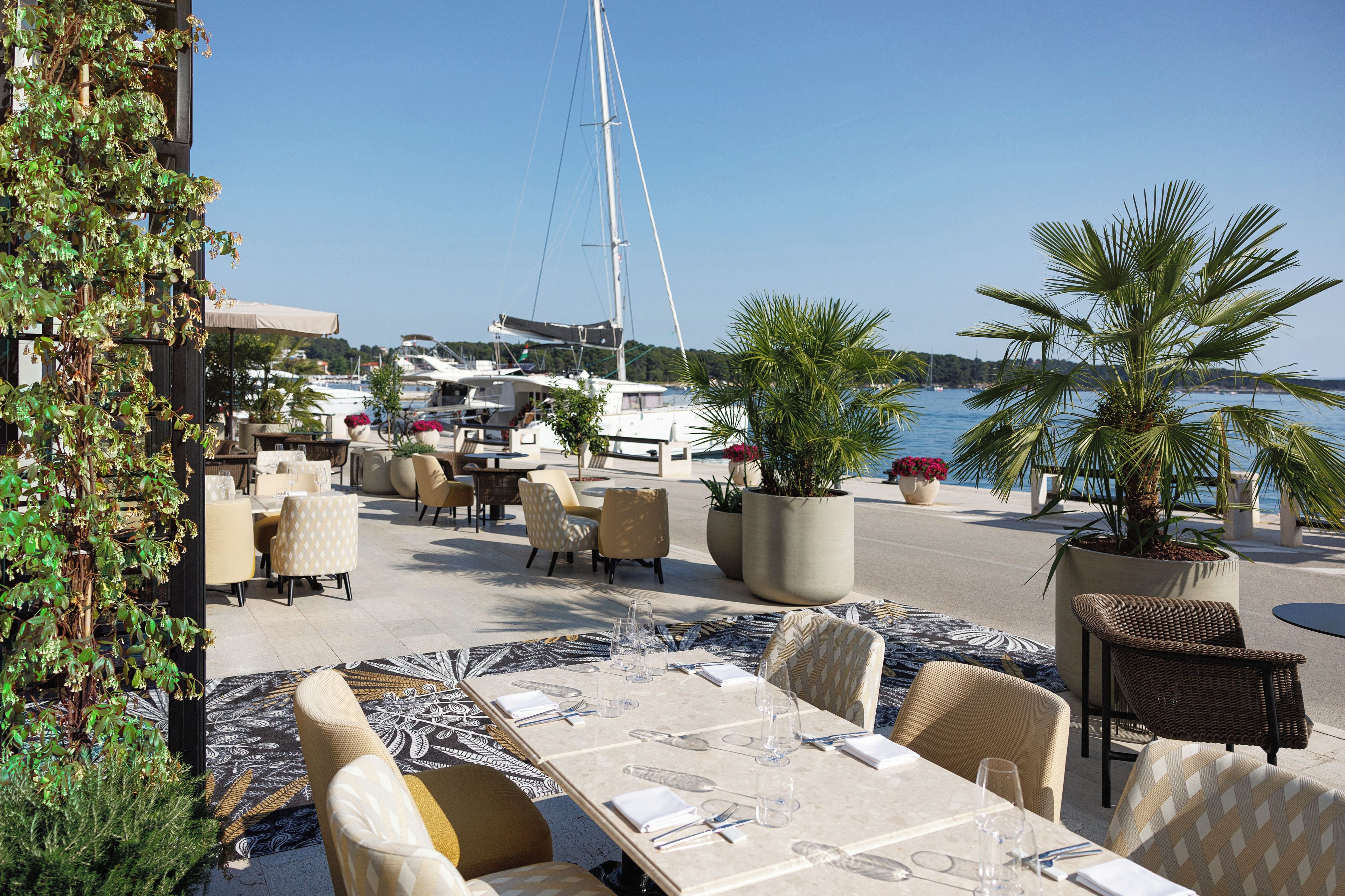 Un espace extérieur aménagé pour les repas, avec des tables et des chaises, des plantes en pot et une vue sur le port de plaisance en arrière-plan.