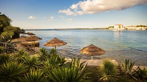 A coastal resort with thatched umbrellas, lounge chairs, and a view of the sea and distant buildings. - Jadran Heritage Hotel Valamar Collection (Poreč)