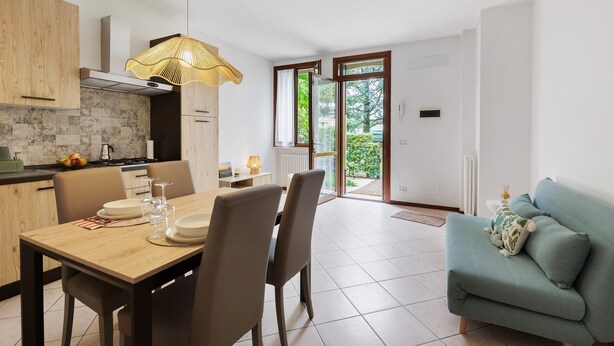 A modern kitchen with a dining area, a sofa, and a view of greenery outside.