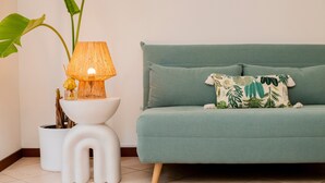 A teal sofa with a patterned pillow, a white side table with a lamp, and a large green plant.