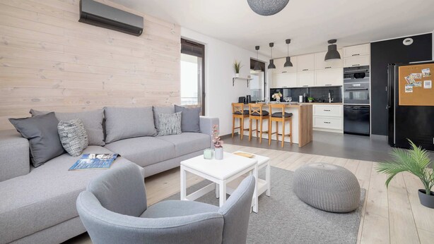 A modern living room with a grey sofa, a white coffee table, and a grey armchair. There is a kitchen in the background with white cabinets and a black refrigerator.