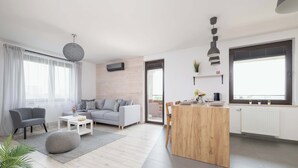 A modern living room with a grey sofa, a white coffee table, and a grey pouf. There's a wooden island with a small table and chairs, and a large window with a view.