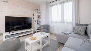 A modern living room with a flat-screen TV, grey sofas, a white coffee table, and a bookshelf.