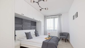 A modern bedroom with a bed, bedside tables, a chair, a window with curtains, and a ceiling light fixture.