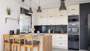 A modern kitchen with white cabinets, a black countertop, and a built-in oven. There are three wooden bar stools with cushions, a potted plant, and a vase with flowers on the counter.
