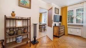 Ein Zimmer mit Holzboden, einer Bücherwand mit unterschiedlichen Gegenständen, einem Fernseher und einem Fenster mit Vorhängen.