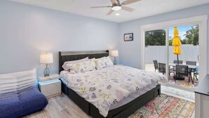 Quarto com cama, mesas de cabeceira, ventilador de teto e vista para um pátio externo com cadeiras e um guarda-sol amarelo.