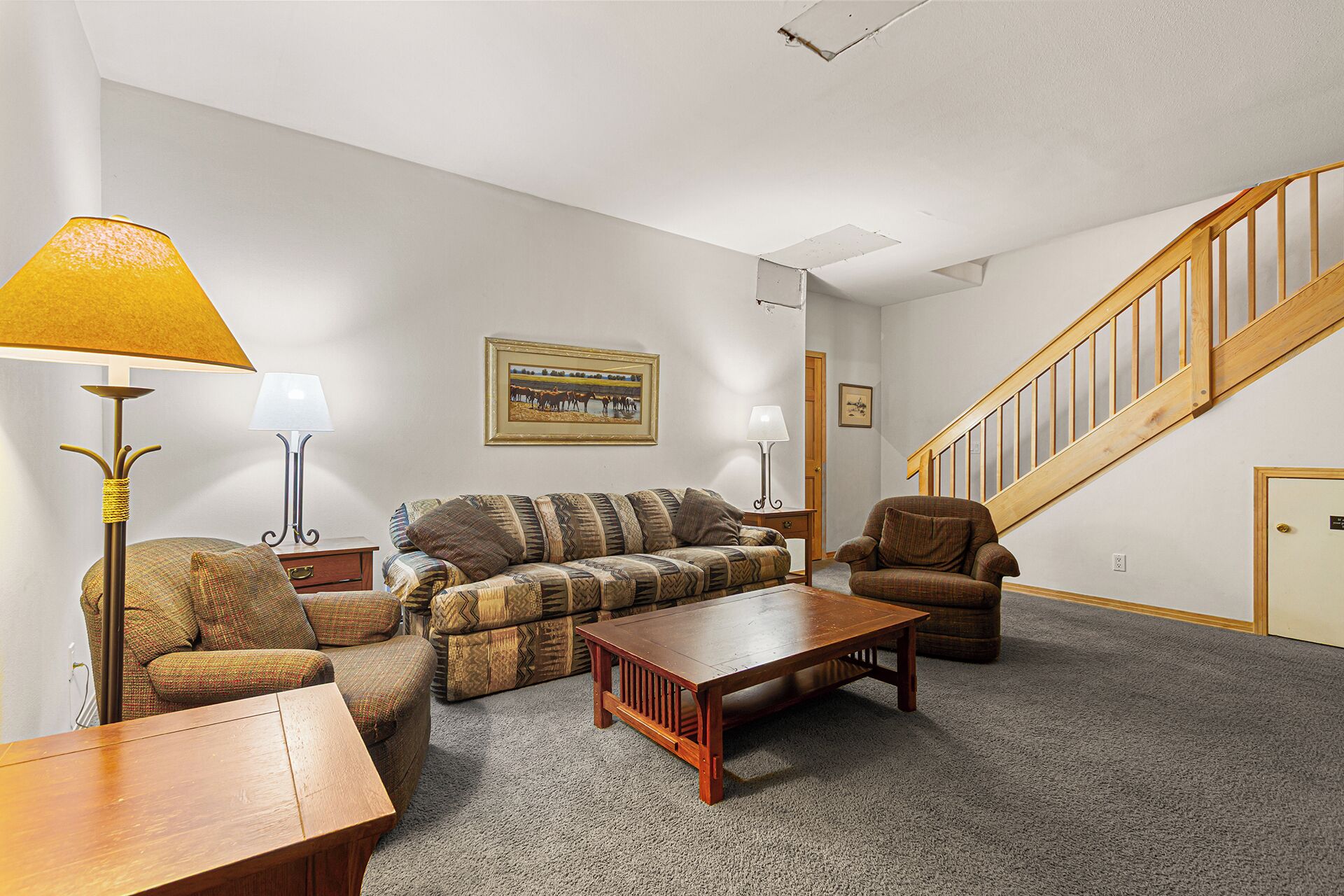 A living room with a sofa, armchair, coffee table, and lamps. There is a staircase with wooden railings and a door.