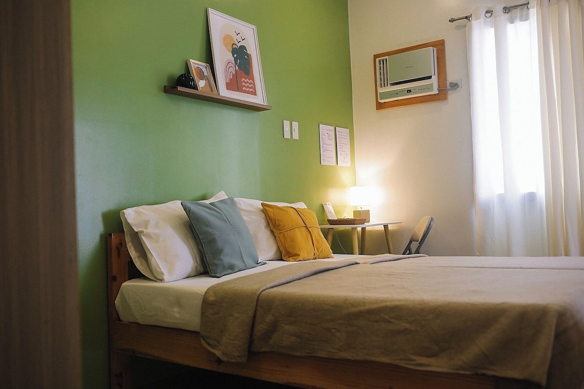 A hotel room with a bed, a nightstand, a lamp, a wall-mounted heater, and a window with curtains.