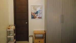 A room with a wooden door, a small wooden cabinet, a shelf with towels, and a framed abstract painting on the wall.