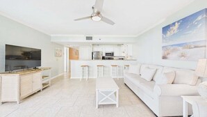 Una sala moderna con televisor, muebles blancos, una cocina al fondo y un ventilador de techo.