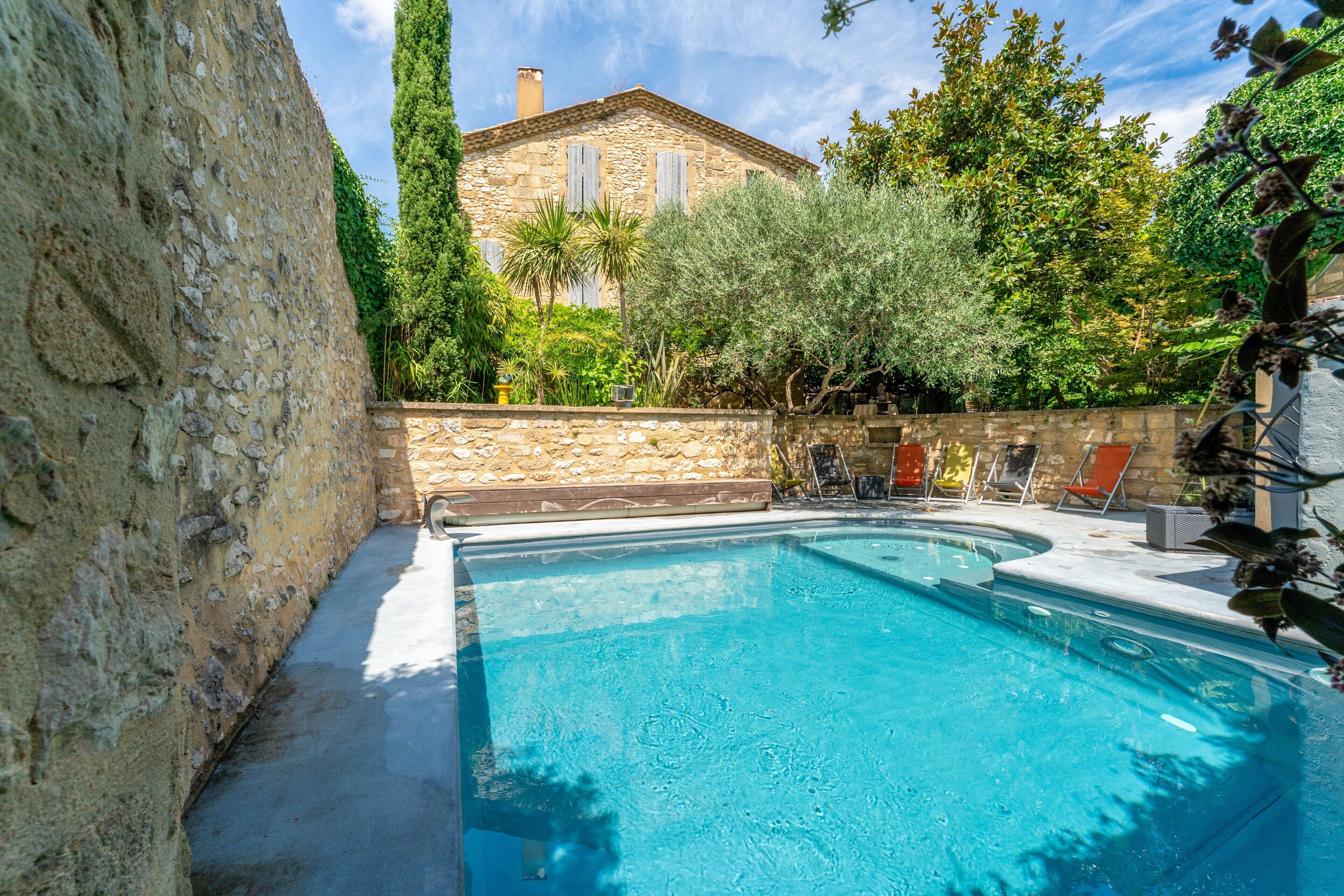Ein Schwimmbecken mit Steinmauer, ein Gebäude mit Fensterläden und ein Garten mit verschiedenen Pflanzen.