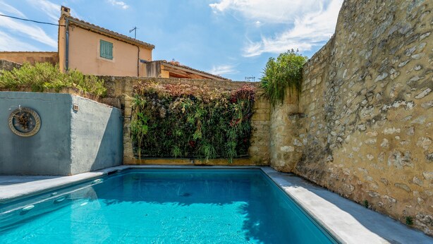 A swimming pool with a stone wall and vegetation on one side, and a building with a tiled roof in the background. - Indigo Room with Shared Pool, Wi-Fi & Air Conditioning (Collias)