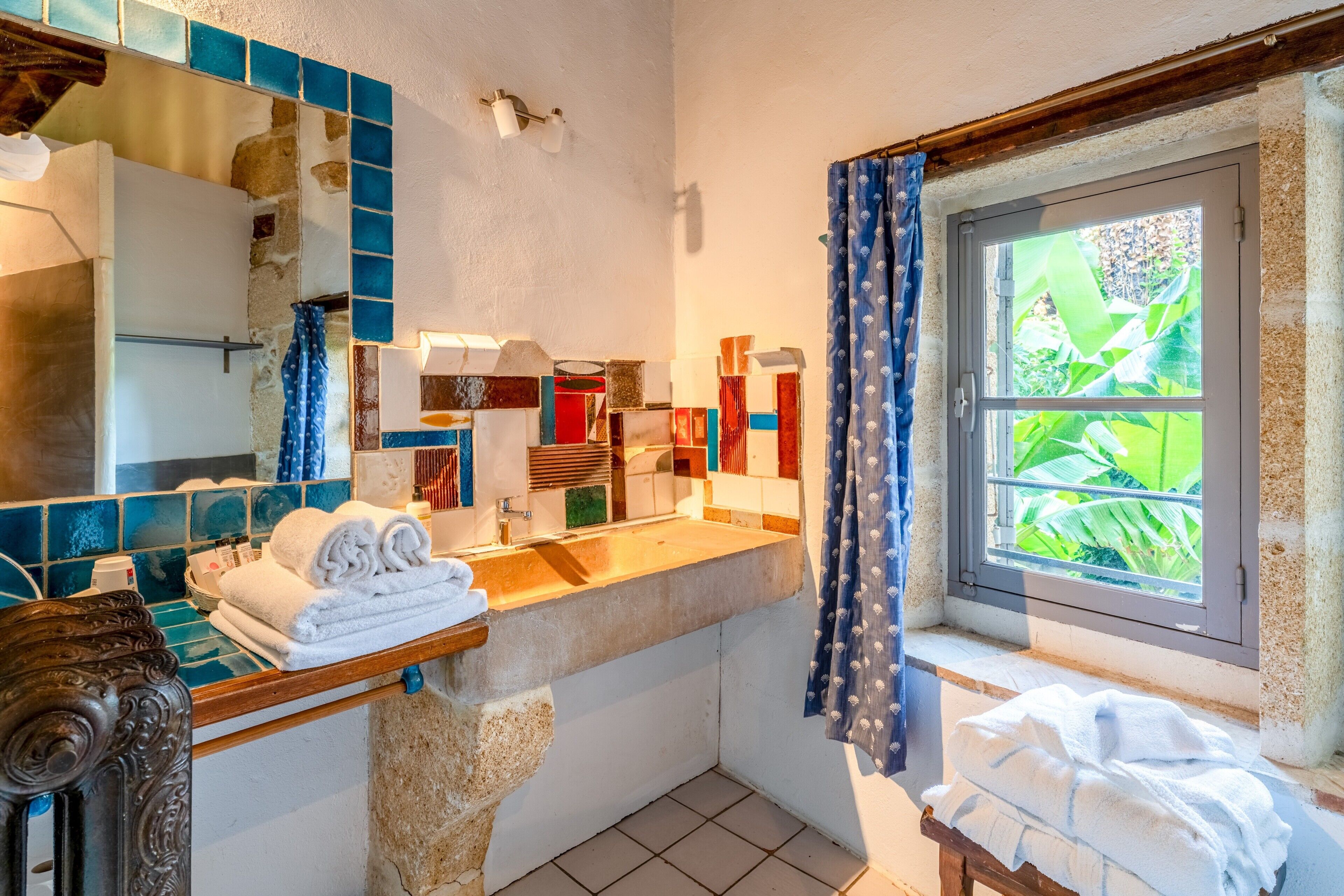 A bathroom with a tiled backsplash, a sink, a mirror, a window with curtains, and towels on the counter.