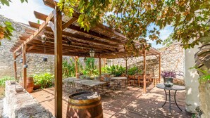 An outdoor patio with wooden tables and chairs, a pergola with hanging lights, and a stone wall backdrop. - Indigo Room with Shared Pool, Wi-Fi & Air Conditioning (Collias)